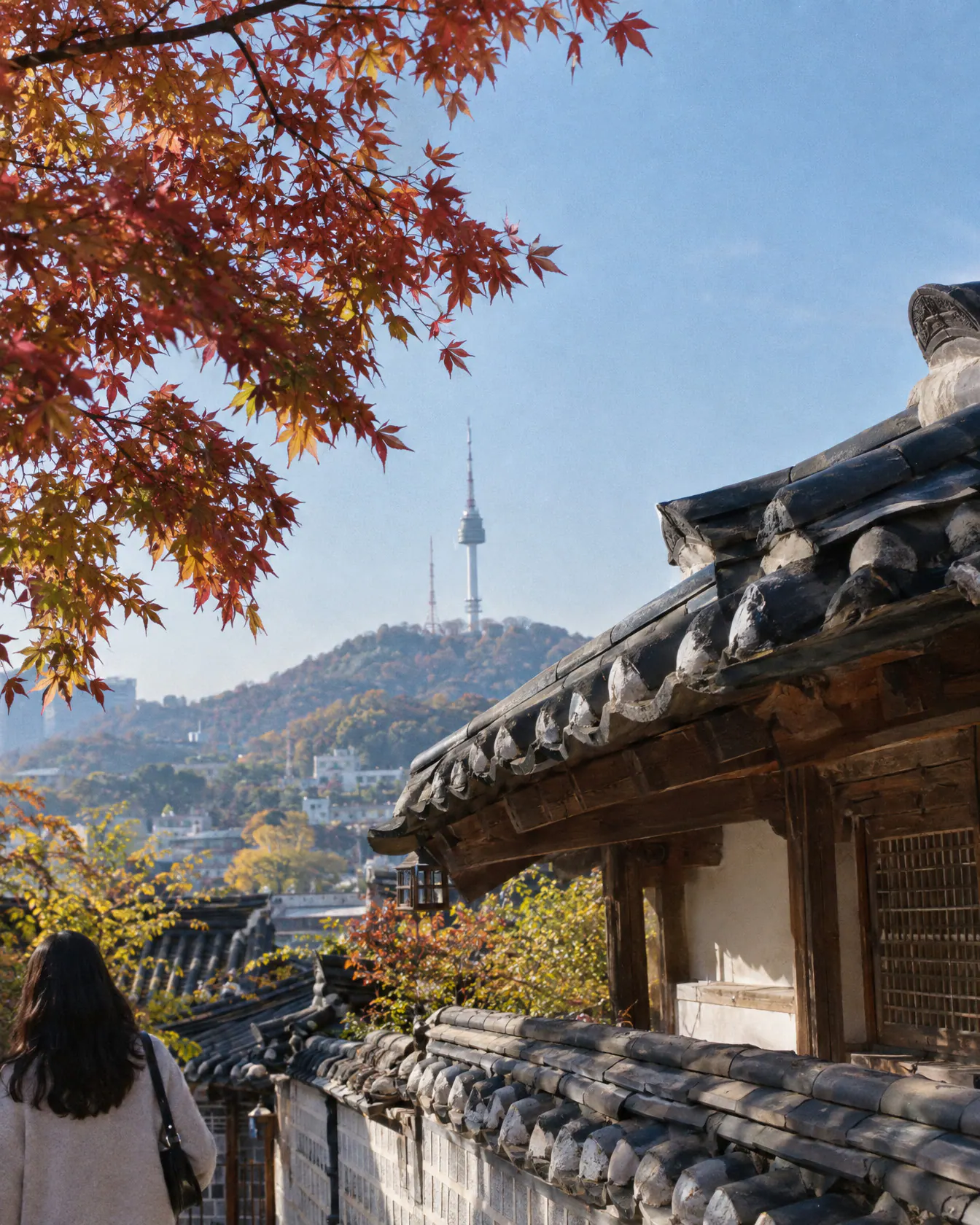 The tiled eaves of a hanok roof in Seoul with a maple branch and a tower beyond